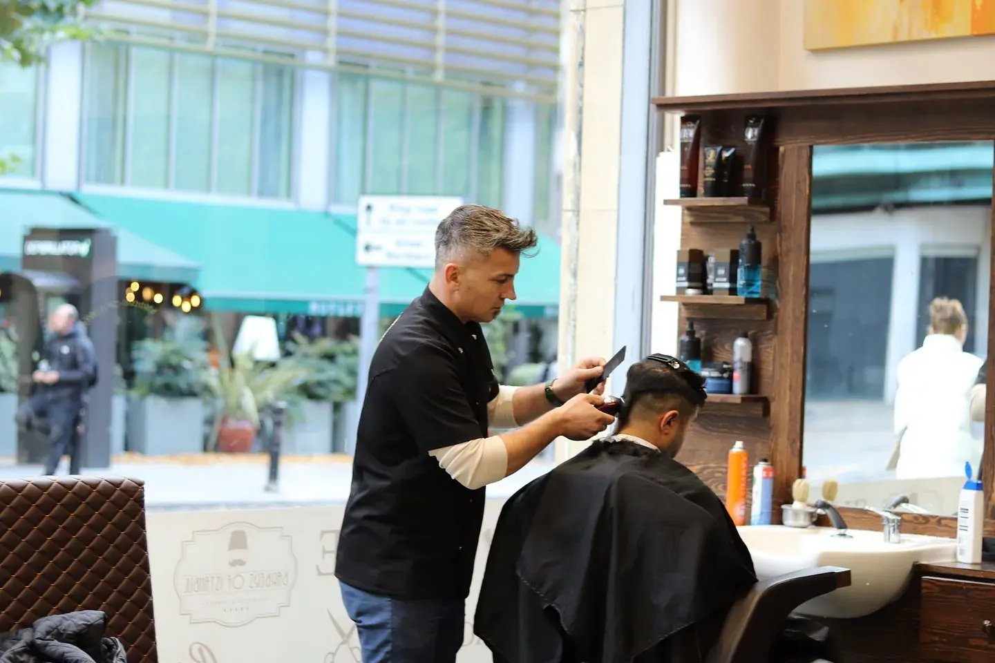 A barber cutting a customer's hair by the shop window on John Dalton Street